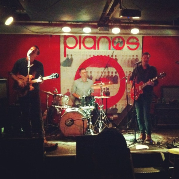 Drei Männer auf der Bühne bei einem Konzert: einer spielt Schlagzeug in der Mitte, und zwei spielen Gitarre und singen auf beiden Seiten, mit einem Banner an der Wand und Lampen, die von der Decke hängen.