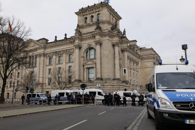 Eine Gruppe von Polizisten steht vor dem Reichstaggeb√§ude in Berlin, Deutschland, mit Fahrzeugen, einem Zaun, Verkehrsampeln, Laternenm√§sten, B√§umen und Flaggen, unter einem klaren Himmel.