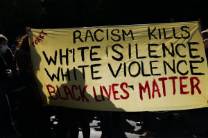 Gruppe maskierter Menschen mit einem Banner "Rassismus tötet Weißes Schweigen, weiße Gewalt, Black Lives Matter" auf einer dunklen Straße.