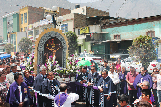 Menschen stehen auf einer Straße mit einem Kreuz und Blumensträußen im Vordergrund, Autos bewegen sich im Mittelgrund, und Gebäude, Bäume und Straßenlaternen im Hintergrund.