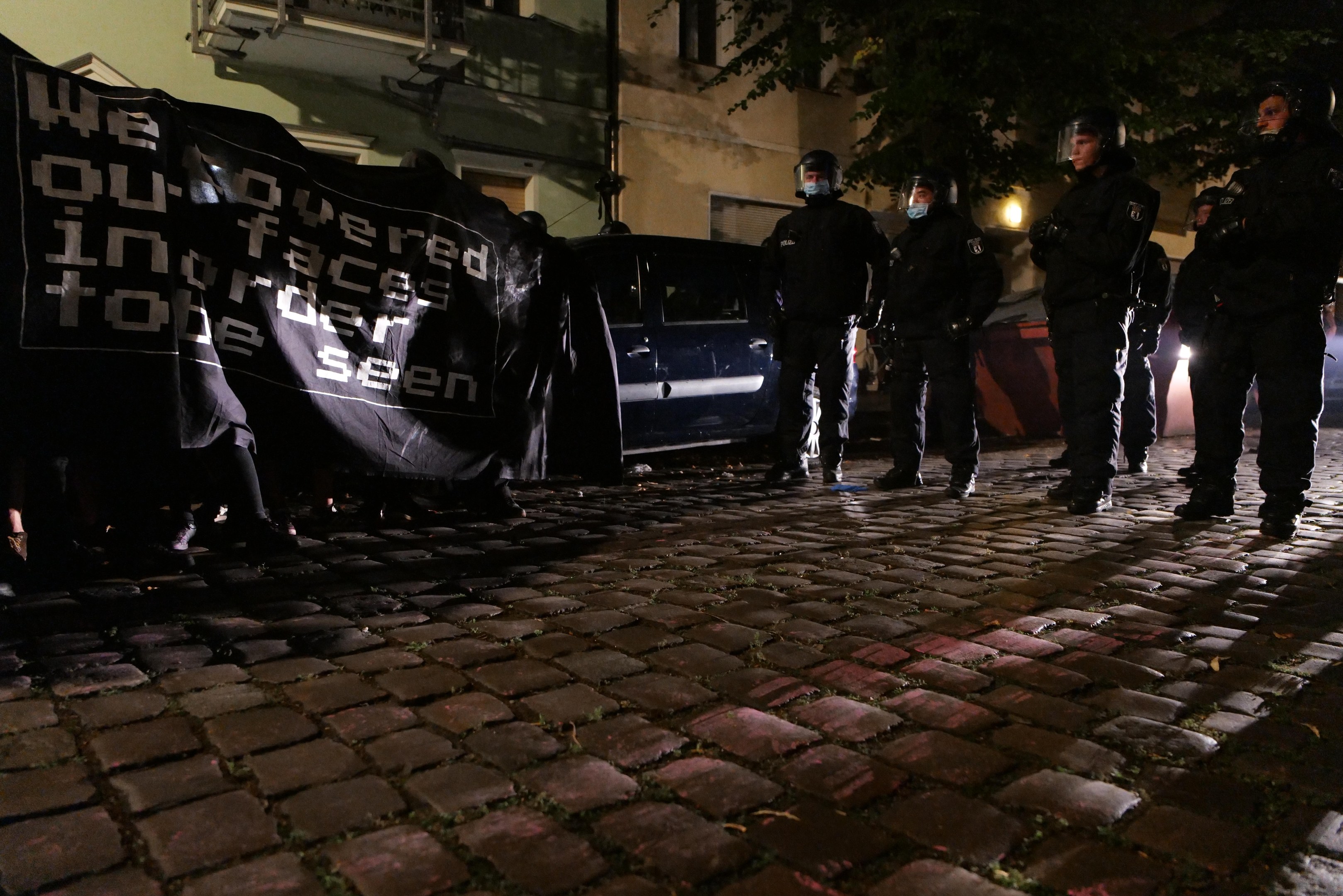 Eine Gruppe von Polizisten in Helmen, die ein Banner auf einem Kopfsteinpflasterweg bei Nacht halten, mit Fahrzeugen, Gebäuden, Bäumen und Straßenbeleuchtung im Hintergrund.