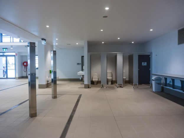 Große öffentliche Toilette mit mehreren Toiletten, tragenden Säulen, einem Mülleimer, einer Tür, einer Tafel, einem Feuerlöscher und Deckenleuchten.