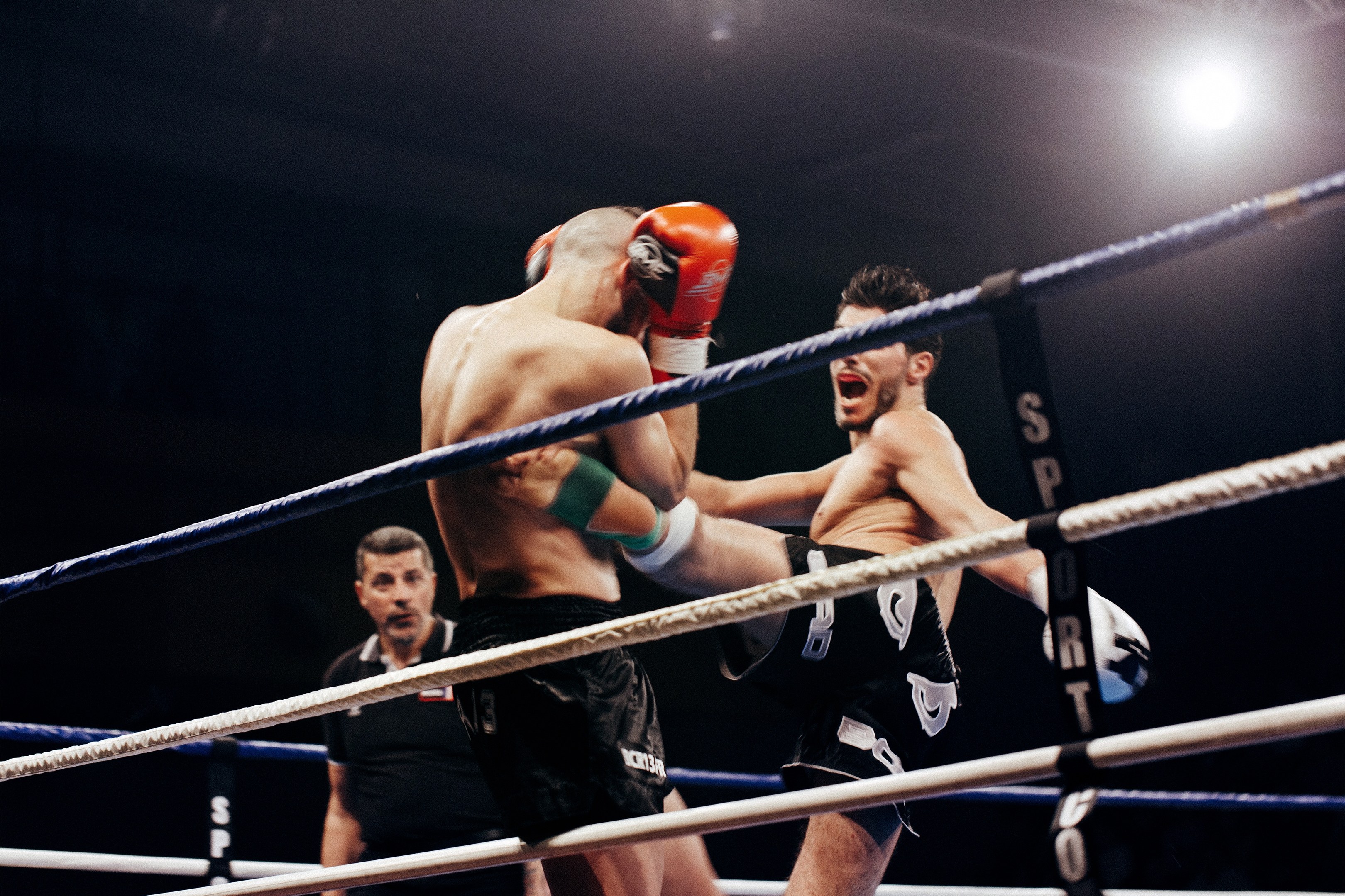 Zwei Boxer im Ring mit einem Referee, der die Aktion beobachtet, unter heller Deckenbeleuchtung.