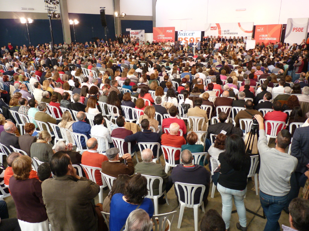 Ein voller Stadion mit vielen sitzenden und stehenden Menschen, eine Bühne mit Lautsprechern auf einem Podium, Banner und Bühnenbeleuchtung.