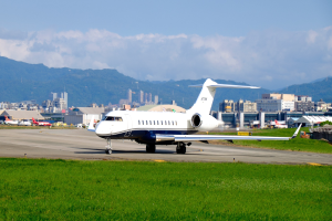 Privater Jet auf einer Flughafenpiste mit saftigem Grün, anderen Flugzeugen, Gebäuden, Bäumen, Bergen und einem klaren blauen Himmel im Hintergrund.