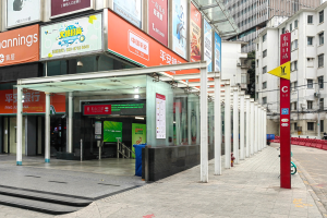 Eine Bushaltestelle auf einer Stadtstraße mit Gebäuden auf beiden Seiten, einer Straße mit einem Müllcontainer in der Mitte, Fußgängern auf dem Gehweg und einer Rolltreppe auf der linken Seite. Rote und gelbe Schilder mit Text sind auf der rechten Seite des Bildes.