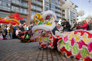 Ein lebendiges chinesisches Neujahrsfest in Amsterdam mit einer Lowen-Tanz-Aufführung vor einer Zuschauermenge, einige halten Kameras, vor dem Hintergrund von Gebauden, Laternenmasten und einem klaren blauen Himmel.