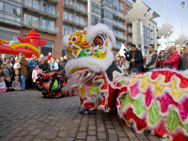 Ein lebendiges chinesisches Neujahrsfest in Amsterdam mit einer Lowen-Tanz-Aufführung vor einer Zuschauermenge, einige halten Kameras, vor dem Hintergrund von Gebauden, Laternenmasten und einem klaren blauen Himmel.