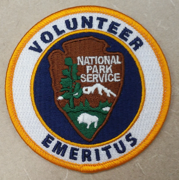 Ein kreisrunder Patch mit einem blauen Rand und einem weißen Hintergrund, der den schwarzen Text 'Volunteer National Park Service Emeritus' in der Mitte eines gelben Kreises zeigt.
