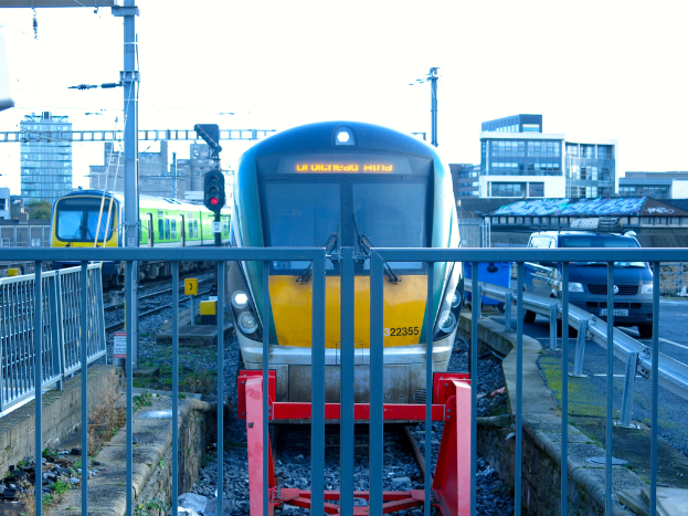 Zwei Züge auf dem Gleis mit einem Zaun im Vordergrund, Pfählen auf dem Land, einer mit einer Signal-Leuchte, einem Auto auf der Straße rechts, Gebäuden im Hintergrund und Himmel oben.