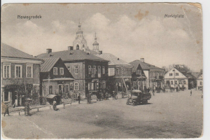 Ein Schwarz-Weiß-Foto einer Stadtstraße in Nowogrodeek, Deutschland, beschriftet mit "Markplatz", das Gebäude, Menschen, Karren, Pfähle, Bäume und Himmel zeigt.