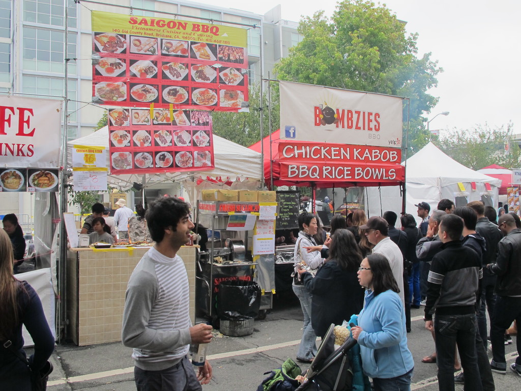 Menschen auf einer Straße außerhalb der Stadt, mit einem Mann in einem grauen und weißen T-Shirt, der eine Wasserflasche hält, und einer Frau in einer blauen Jacke, die einen Kinderwagen schiebt. Im Hintergrund sind Stände mit weißen und roten Zelten, farbige Banner und Gebäude, Bäume und eine Straßenlaterne zu sehen.