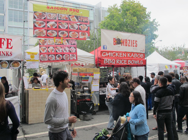 Menschen auf einer Straße außerhalb der Stadt, mit einem Mann in einem grauen und weißen T-Shirt, der eine Wasserflasche hält, und einer Frau in einer blauen Jacke, die einen Kinderwagen schiebt. Im Hintergrund sind Stände mit weißen und roten Zelten, farbige Banner und Gebäude, Bäume und eine Straßenlaterne zu sehen.