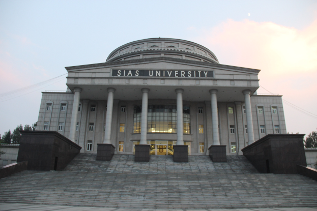 Gro├čer Bau mit S├Ąulen, Fenstern, Türen und Treppen, umgeben von B├Ąumen unter einem klaren blauen Himmel, mit Text auf der Fassade, der wahrscheinlich den Namen einer Universit├Ąt angibt.