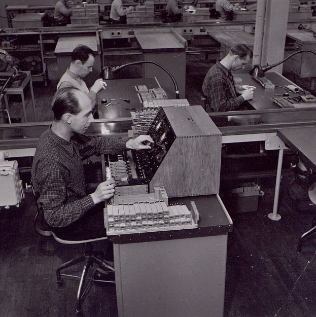 Schwarzes und weißes Bild von Männern, die an Tischen arbeiten, die Computer in einer Fabrikumgebung haben.