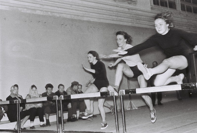Zwei Frauen in der Luft, die über ein Hindernis in einem Turnsaal springen, mit Zuschauern links und Fenstern an der Hintergrundwand.