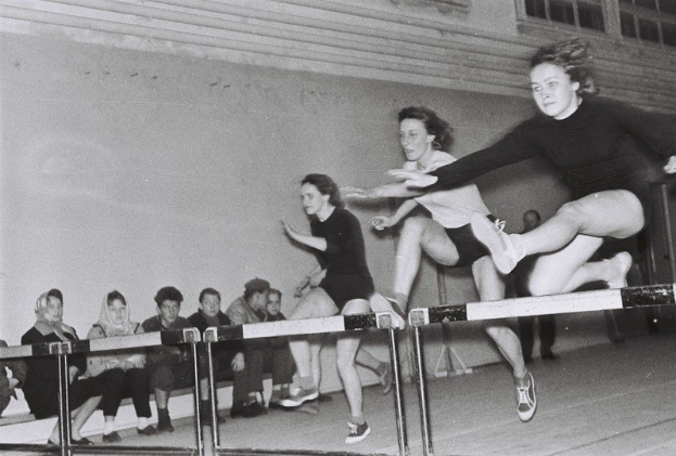 Zwei Frauen in der Luft, die über ein Hindernis in einem Turnsaal springen, mit Zuschauern links und Fenstern an der Hintergrundwand.