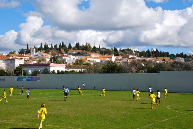 Eine Gruppe von Menschen spielt ein Spiel auf einem Feld mit Plakaten an einer nahen Wand, Bäumen, Pfählen, Häusern und Wolken im Hintergrund.