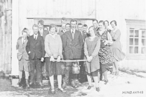 Schwarzes und weißes Foto von Menschen, die lächeln und ein Band halten, vor einem Gebäude mit einem Fenster und einer Tür, das die Eröffnung einer neuen Schule feiert.