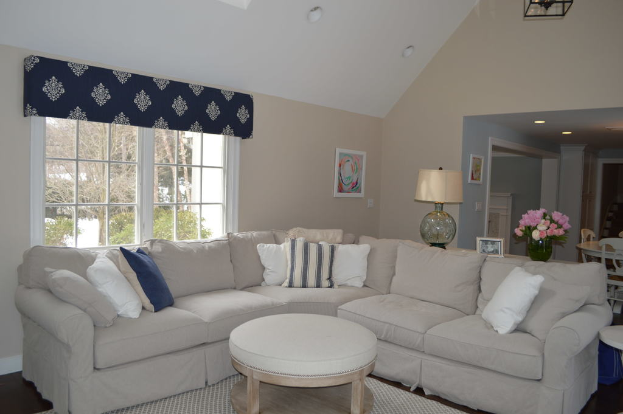 Ein Wohnzimmer mit einer Couch, Kissen, einem Tisch, Stühlen, einem Blumenstrauß, einem Foto Rahmen, einer Alarm Uhr und einem Fenster mit Deckenleuchten.