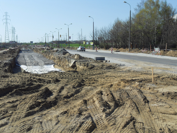 Baustelle mit einem großen Haufen Erde neben der Straße, Fahrzeuge in Bewegung, Laternenpfähle, Strommasten mit Drähten, Bäume und einen klaren blauen Himmel.