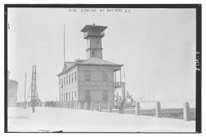 Schwarz-weiß-Foto einer Feuerwache in Battery, New York, mit einem Gebäude, Menschen, Pfählen, einem Zaun, Booten auf dem Wasser und einem bewölkten Himmel.