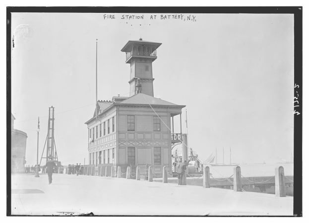 Schwarz-weiß-Foto einer Feuerwache in Battery, New York, mit einem Gebäude, Menschen, Pfählen, einem Zaun, Booten auf dem Wasser und einem bewölkten Himmel.