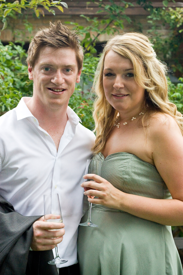 Ein Paar lächelt und hält Weingläser in der Hand, mit ein paar Bäumen im Hintergrund.