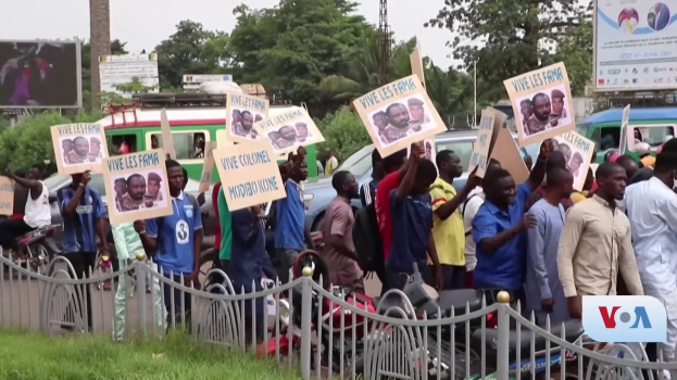 Eine Gruppe von Menschen h├Ąlt Schilder in H├Ąnden und steht vor einem Zaun, umgeben von Fahrzeugen, B├Ąumen, Pfosten und Plakatw├Ąnden, mit einem Bildschirm und Gras im Hintergrund, wahrscheinlich protestieren sie gegen eine Regierungsentscheidung, die Video-Konferenzen verbietet.