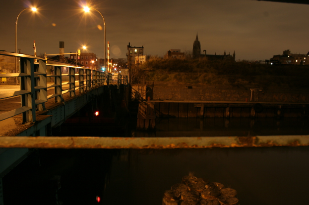 Eine Brücke überspannt einen Fluss mit zahlreichen Straßenlaternen auf ihrer Länge und zahlreichen Gebäuden im Hintergrund.
