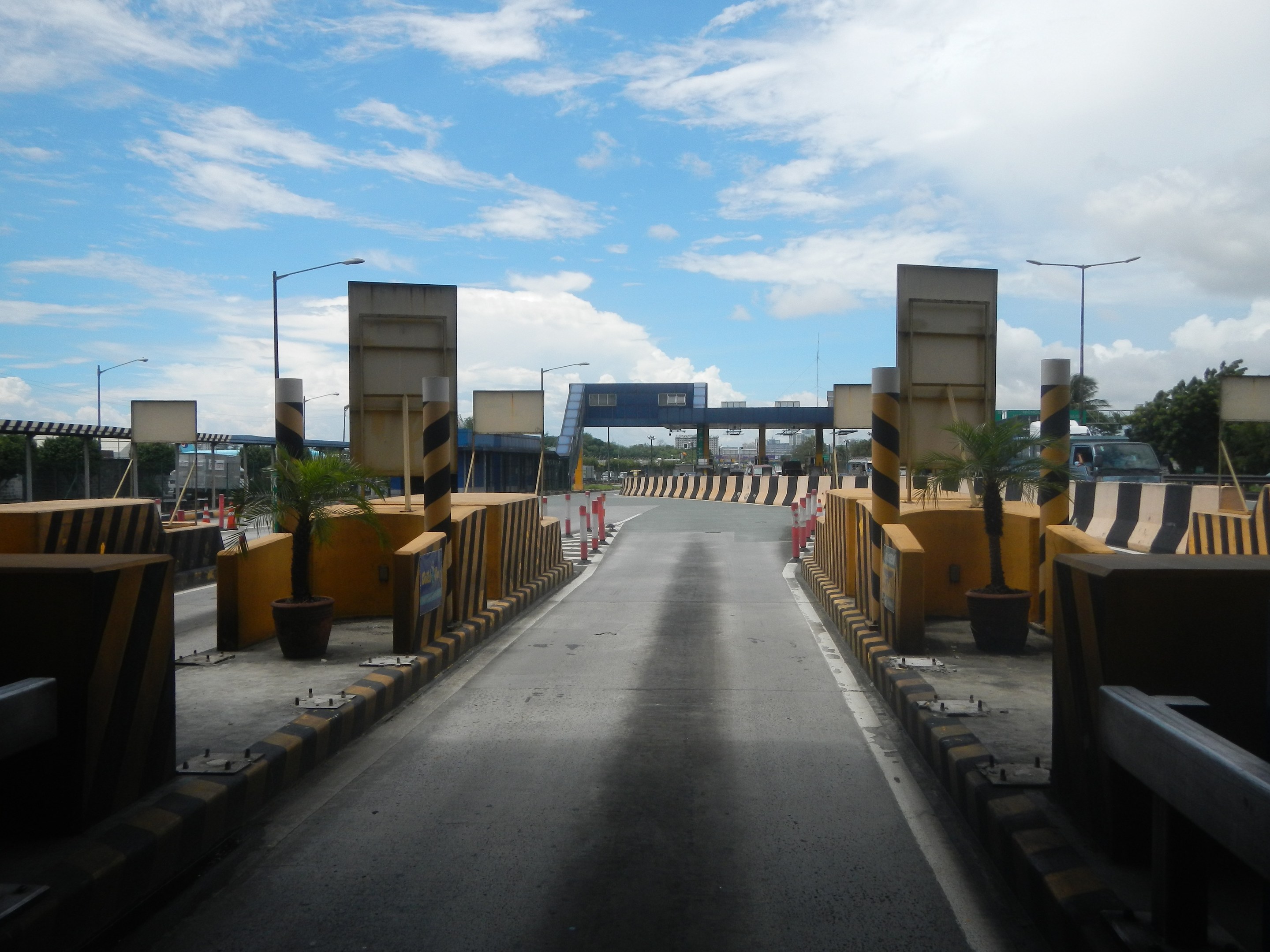 Straße mit einer Mautstelle, Absperrpflöcke, Verkehrskegel, Topfpflanzen, Hinweistafeln, Straßenmasten, Bäume, und eine Brücke, sowie einen bewölkten Himmel.