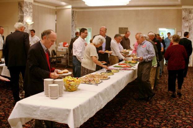Eine Gruppe von Menschen mit Tellern in der Hand, die um einen Tisch mit Essen, Schalen und L├Âffeln versammelt sind, mit rotem Teppich und Säulen im Hintergrund.