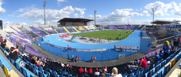 Ein großes Stadion voller Zuschauer, die einer Leichtathletikveranstaltung zusehen, mit Flutlicht, Hütten, Bäumen und einem bewölkten Himmel im Hintergrund.