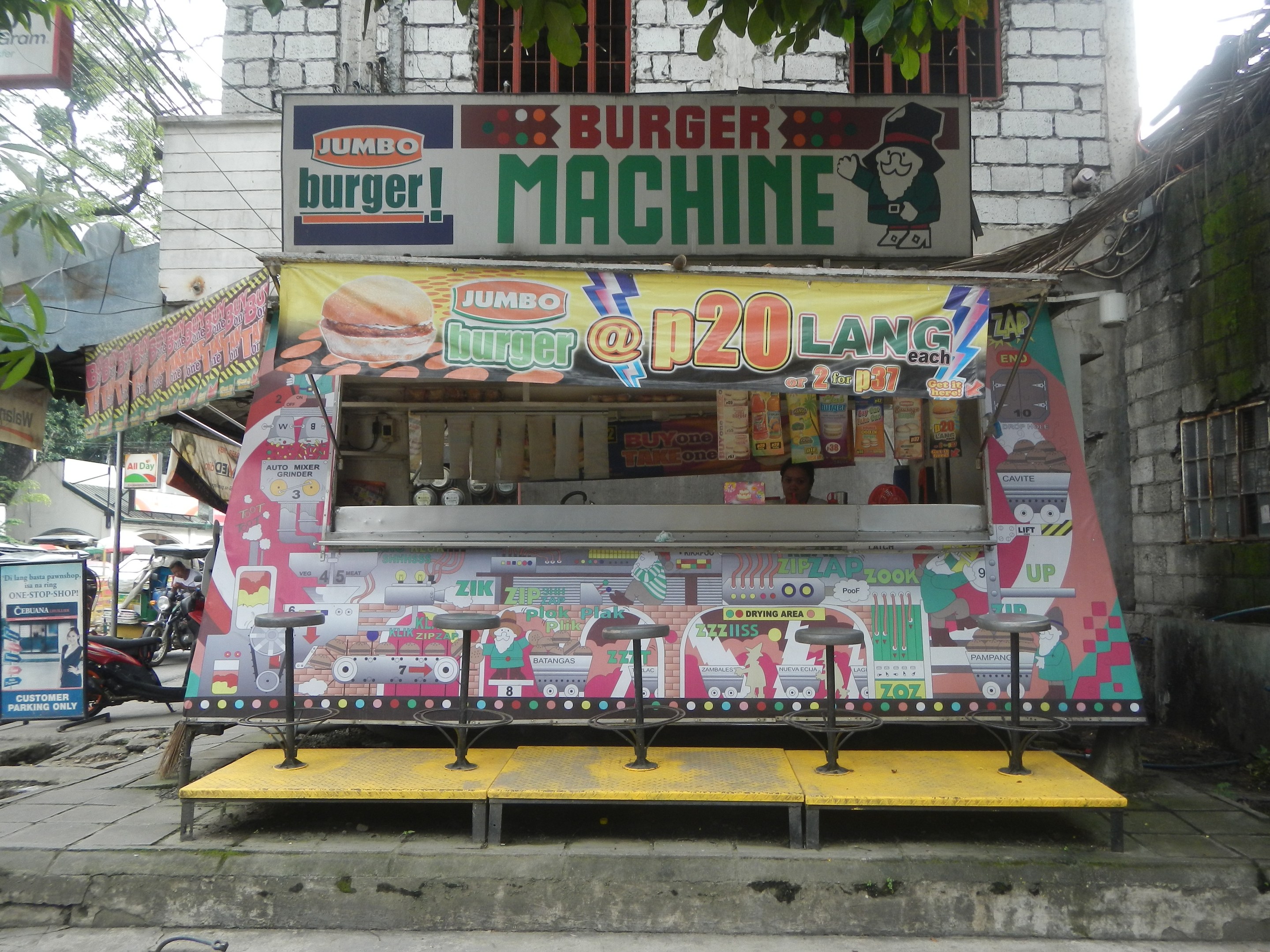 Ein leuchtend gelber und roter Foodtruck in Form einer Jumbo-Burger-Maschine parkt auf einer städtischen Straße, umgeben von Gebäuden, Bäumen, Menschen und Fahrzeugen.