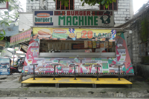 Ein leuchtend gelber und roter Foodtruck in Form einer Jumbo-Burger-Maschine parkt auf einer städtischen Straße, umgeben von Gebäuden, Bäumen, Menschen und Fahrzeugen.