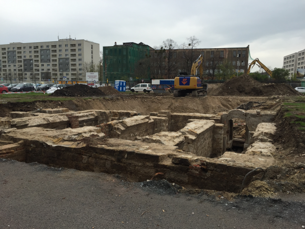 Eine Baustelle in Berlin mit einem großen Haufen Erde, Baggern, Fahrzeugen und Gebäuden mit Fenstern im Hintergrund, unter einem bewölkten Himmel.