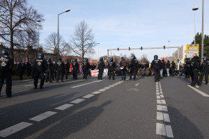 Gruppe von Polizisten in schwarzen Uniformen und Helmen, die an der Straße mit Laternenmasten, Ampeln, Bäumen, Gebäuden und einem klaren blauen Himmel im Hintergrund stehen.