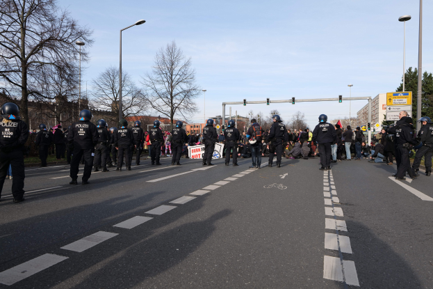 Gruppe von Polizisten in schwarzen Uniformen und Helmen, die an der Straße mit Laternenmasten, Ampeln, Bäumen, Gebäuden und einem klaren blauen Himmel im Hintergrund stehen.