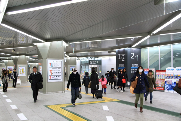 Menschen mit Gepäck durch einen Bahnhof gehen, überirdische Schilder und Deckenleuchten.