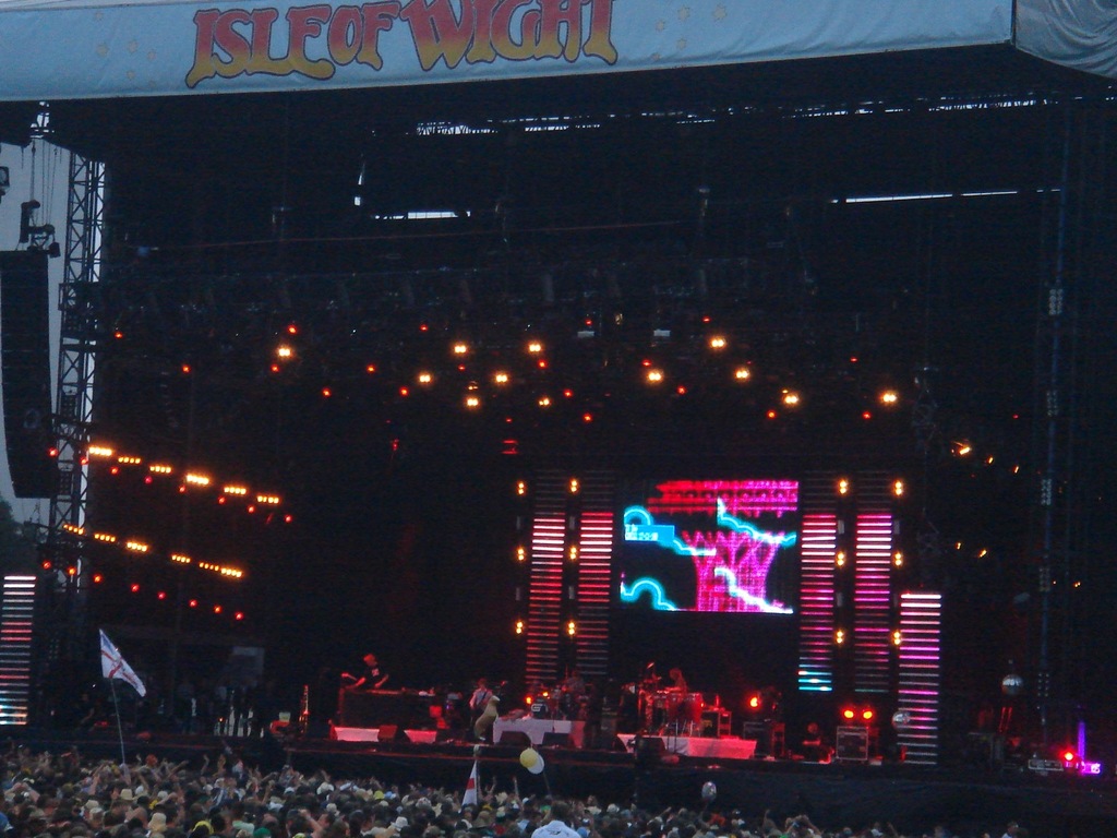 Eine Bühne mit Lichtern, einem Bildschirm und Musikern, die Instrumente spielen, mit einem Banner oben, einer Menge unten und Fahnen mit Stangen und Ballons drumherum.