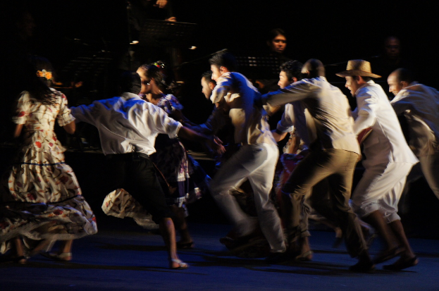 Eine Gruppe von Menschen tanzt im Vordergrund mit einer anderen Gruppe, Stühle und Mikrofone im Hintergrund, alles auf einem Boden.