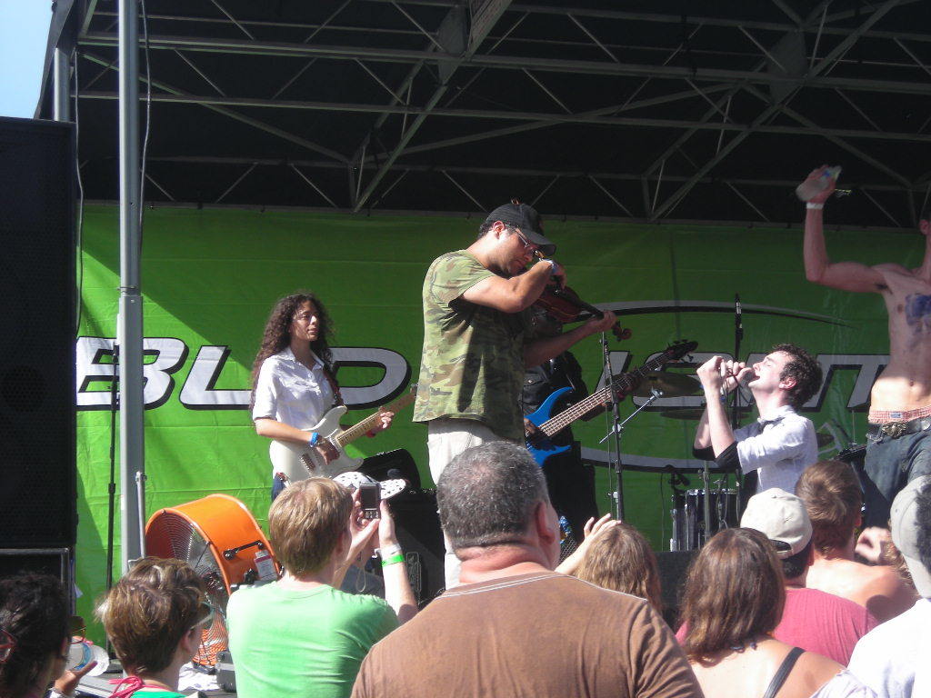 Eine Gruppe von Musikern, die auf der Bühne mit verschiedenen Instrumenten spielen, ein großer schwarzer Lautsprecher links und ein Publikum, das vor einem Podium mit einem grünen Banner steht.