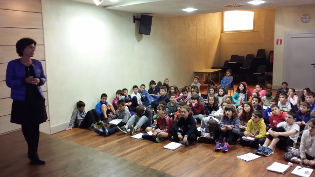 Eine Gruppe von Kindern, Jungen und Mädchen, sitzt auf dem Boden mit Büchern vor ihnen, während eine Frau auf einer Bühne links steht.