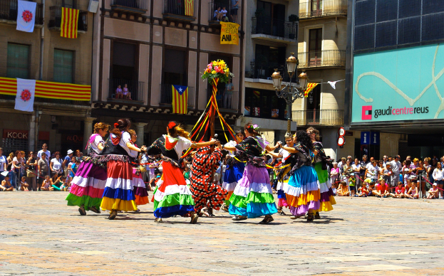 Eine Gruppe Frauen in bunten Kleidern tanzt auf der Straße, umgeben von einer Menge Menschen, mit Gebäuden, Gelöndern, Balkonen, Fahnen, einem Laternenmast und einer Tafel mit Aufschrift im Hintergrund.