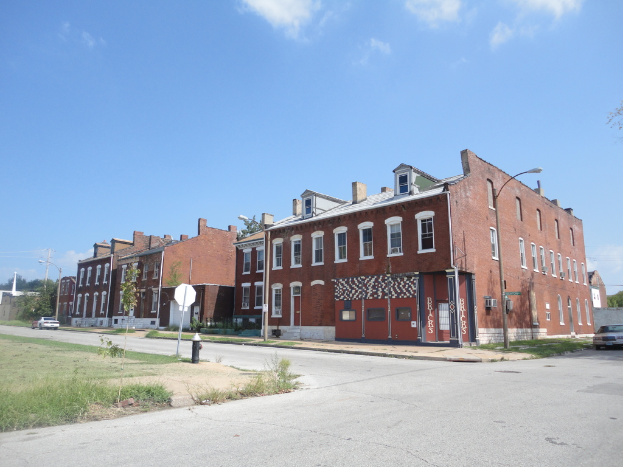 Ein Gebäude mit Straßenlaternen, Masten und Bäumen, mit Fahrzeugen auf beiden Seiten, einem Gehweg und Gras am Boden und einem Himmel oben.