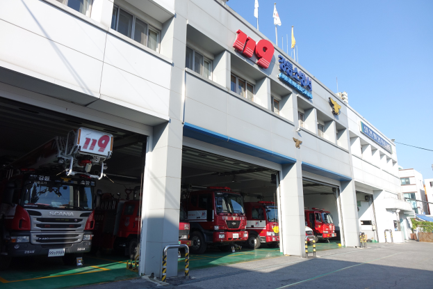 Feuerwache mit mehreren Feuerwehrautos vor dem Gebäude mit Fenstern, Säulen, Namensschildern und Flaggen im Vordergrund, vor anderen Gebäuden und einem klaren blauen Himmel.