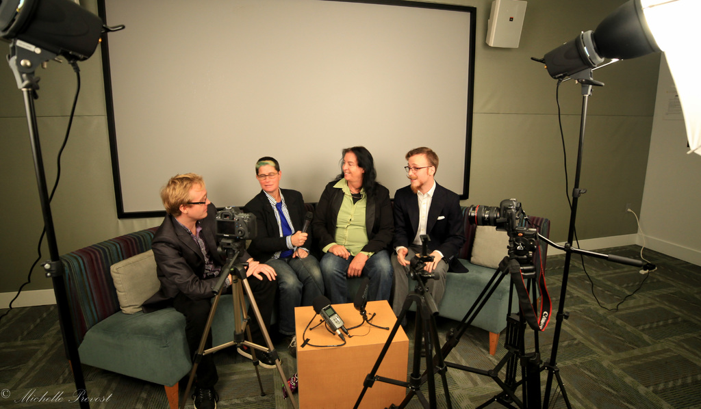 Eine Gruppe von drei Personen sitzt auf einem Sofa und gibt ein Interview, wobei eine Person in einem schwarzen Mantel und Hose links, eine Person in einem Mantel und blauer Krawatte in der Mitte ein Mikrofon hält und eine Person rechts lächelt. Ein Mikrofonrekorder befindet sich auf dem Tisch in der Mitte, Kameras sind auf der rechten und linken Seite und große Scheinwerferständer sind vorhanden, mit einem Projektionsschirm im Hintergrund.