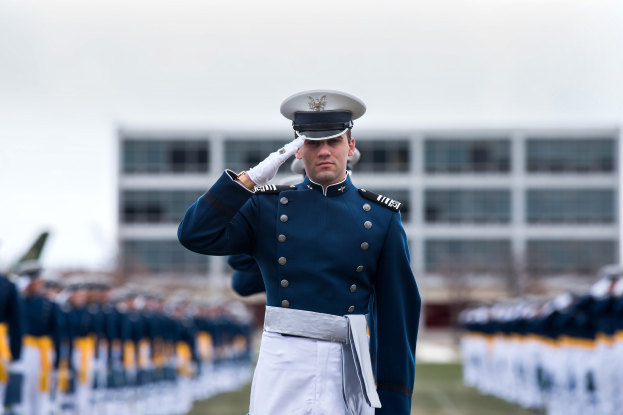 Ein Mann in militärischer Uniform salutiert auf einer Abschlussfeier umgeben von einer Menge, mit einem Gebäude und dem Himmel im Hintergrund.