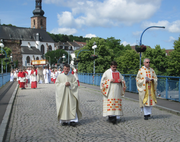 Eine Gruppe von Priestern, die eine Straße mit Laternen, Geländern und Bäumen entlanggehen; im Hintergrund sind Gebäude und ein bewölkter Himmel zu sehen; einer der Priester hält ein Buch.