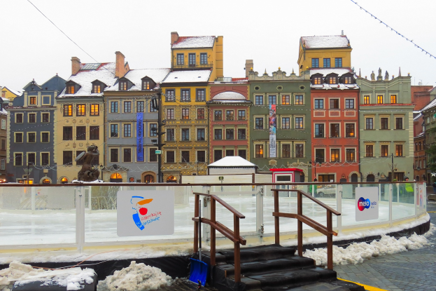Städtisches Eislaufstadion in einem Stadtplatz mit schneebedecktem Boden, umgeben von Gebäuden, Treppen, Laternenmasten und Bäumen unter einem Himmel.